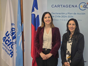 Dos mujeres posan para la cámara al lado de banderas de Chile, Naciones Unidas y ACNUR. A sus espaldas un rótulo mencional el Plan de Chile y la campaña Cartagena+40.