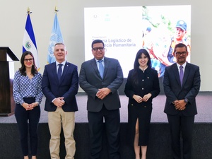 Tres hombres y dos mujeres con vestimenta formal, de pie frente a un foro y dos banderas al fondo