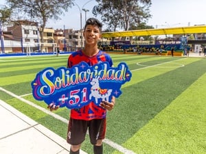 Un joven que viste uniforme de fútbol rojo, sostiene un letrero colorido en el que se lee SOLIDARIDAD +51 en una cancha de fútbol