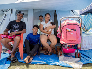 Una familia de cinco miembros, entre ellos un adolescente, un niño y una bebé, está sentada frente a la entrada de una tienda de campaña