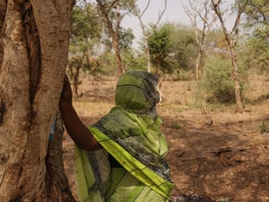 Una mujer recargada en un árbol tiene la mirada puesta en unos matorrales