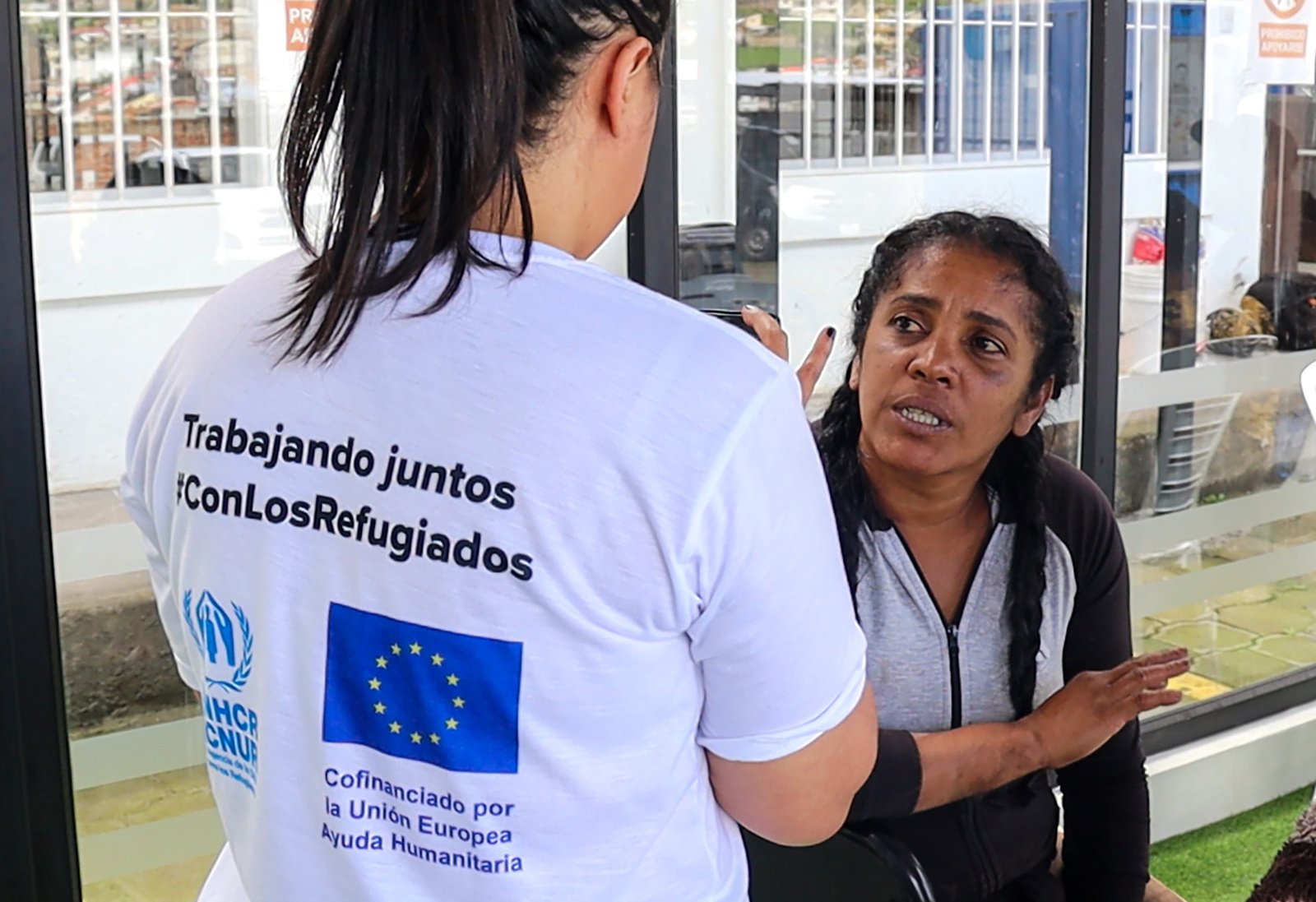 Una mujer con una camisa con los logos de ACNUR y ECHO, de espaldas, hablas con otra mujer que está sentada frente a ella.
