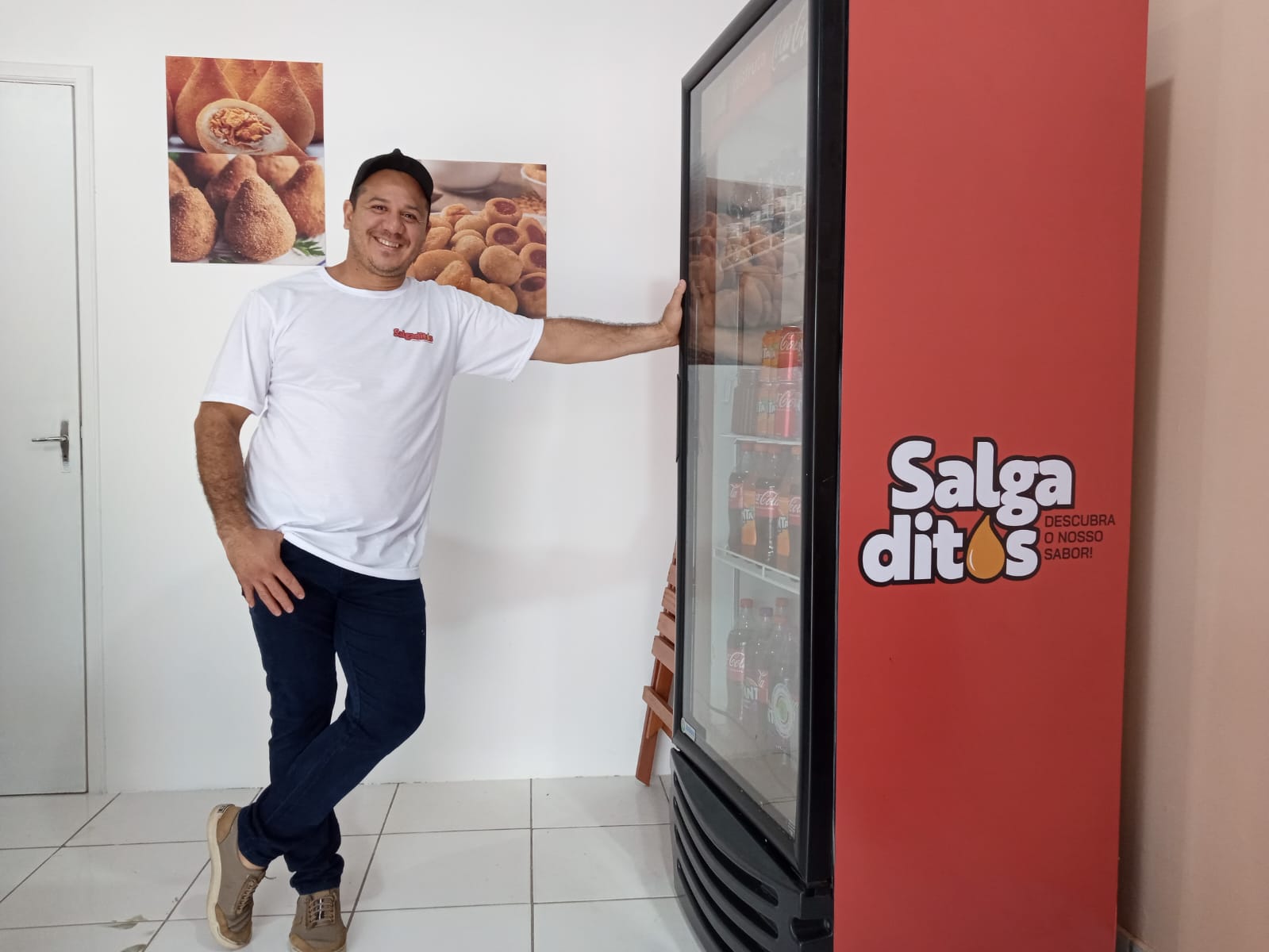Um homem vestindo uma camiseta branca e calças escuras está de pé com uma mão apoiada em uma máquina de venda automática vermelha. A máquina tem o texto &quot;Salgaditos: Descubra o Melhor Sabor&quot; escrito nela. Há dois cartazes na parede atrás dele, ambos exibindo imagens de salgados. O chão é de azulejos e há uma porta fechada ao fundo.
