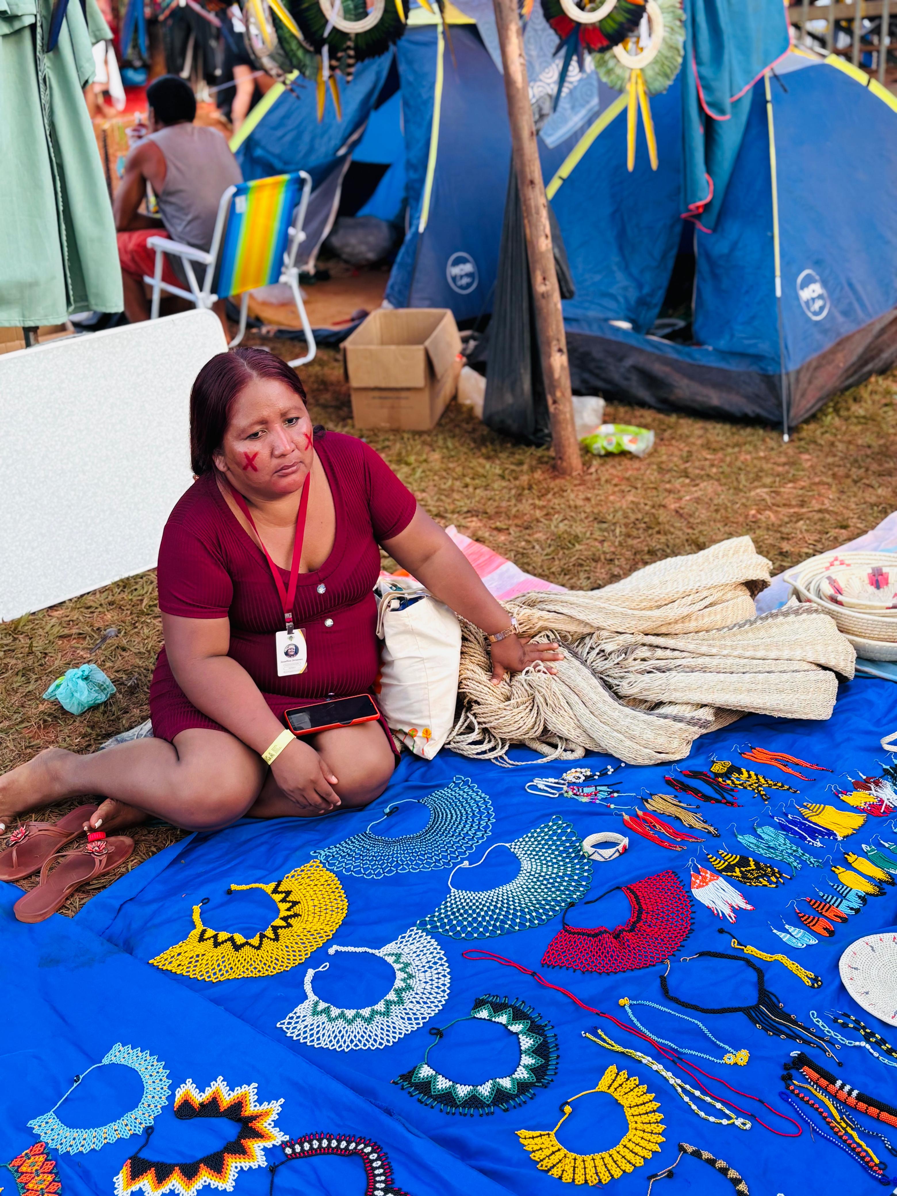Indígena Warao expõe artesanatos no Acampamento Terra Livre 2025 Mulher indígena da etnia Warao está sentada em frente a uma barraca de camping, exibindo colares artesanais coloridos dispostos sobre um tecido azul no chão. Ao fundo, há outras barracas e pessoas.