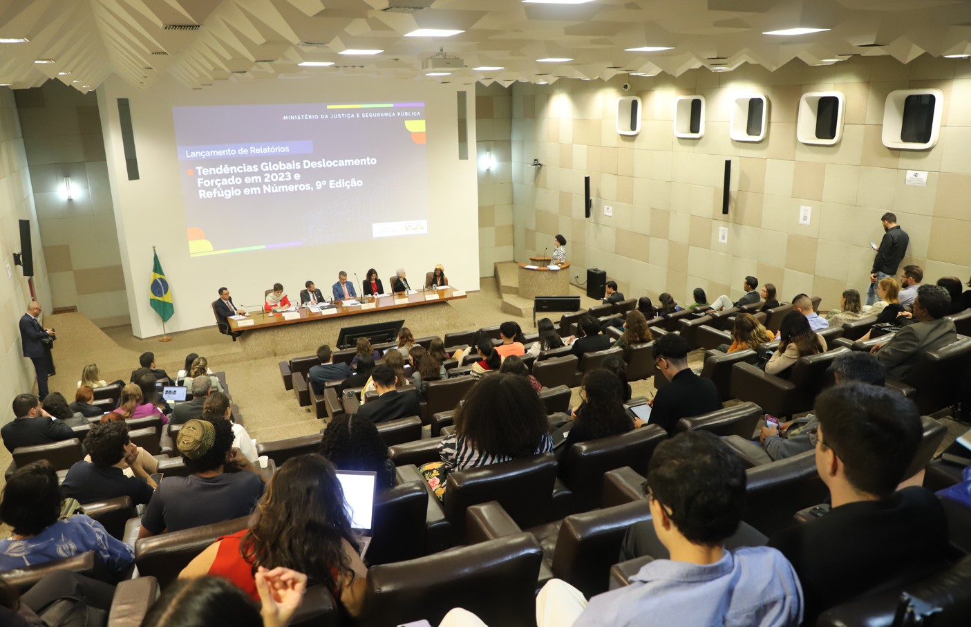 Pessoas em um auditório assistindo a outras pessoas em uma mesa com uma grande tela ao fundo.
