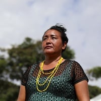 Alida usa colares de contas amarelas posa ao ar livre durante o dia. A imagem foi feita em ângulo baixo, destacando a pessoa contra o céu parcialmente nublado. Ao fundo, aparecem árvores desfocadas e parte de uma tenda, criando um contexto de evento realizado em área aberta.