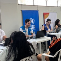 Pessoas atendem solicitantes em uma sala com mesas, computadores e cadeiras durante ação de atualização documental. Ao fundo, banners do ACNUR e do Serviço Jesuíta a Migrantes e Refugiados (SJMR) estão posicionados atrás da equipe de atendimento.