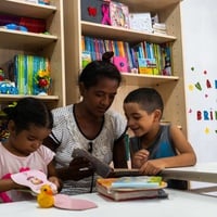 Crianças e uma pessoa adulta folheiam livros em um espaço de leitura com estantes repletas de materiais educativos e brinquedos.