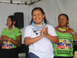 Três pessoas de pé, todas com colares coloridos. A pessoa no centro está usando uma camiseta branca com o texto 'MOBILIS' e um mapa do mundo. As duas outras pessoas estão usando camisetas verdes com a imagem de Nossa Senhora de Coromoto e o texto 'Nossa mãe Coromoto Patrona de Venezuela'