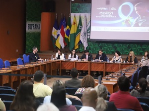 Evento oficial com várias pessoas sentadas em uma mesa longa à frente de um auditório. Atrás dos palestrantes, há bandeiras do Brasil e de estados brasileiros. Um telão exibe a frase 'resiliência climática e inclusão socioeconômica'. A palavra 'SUFRAMA' aparece à esquerda, indicando relação com a Superintendência da Zona Franca de Manaus.