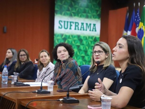 Painel com seis mulheres sentadas à mesa com microfones, participando de uma conferência. Ao fundo, há uma faixa verde e branca com o texto 'SUFRAMA'. Sobre a mesa, há garrafas de água e copos.