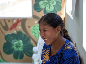mulher indígena de perfil com camiseta azul e pinturas de flores verdes ao fundo.
