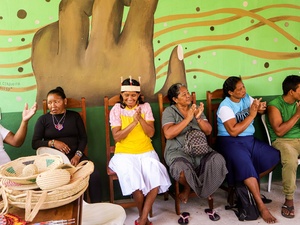 Grupo de pessoas indígenas sentadas em uma fileira de cadeiras, algumas aplaudindo e sorrindo, em frente a um mural colorido com arte representando uma mão em destaque e elementos gráficos abstratos. Cestos artesanais indígenas estão sobre uma mesa à esquerda.