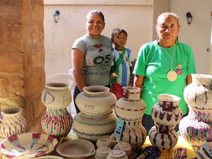 Três pessoas indígenas em pé, sendo duas mulheres e uma criança ao meio, atrás de uma mesa com cestos e itens artesanais de diferentes formas, tamanhos e cores, exibindo padrões detalhados. 