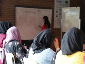 Sala de aula com estudantes sentadas, usando lenços na cabeça, voltadas para um quadro branco onde uma pessoa instrutora escreve ou explica algo. Ao lado do quadro há um flip chart, e ao fundo, uma parede de tijolos com cartazes.