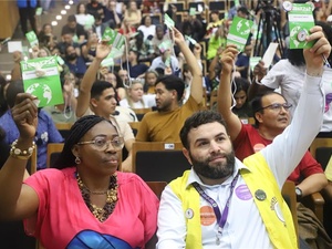 Auditório cheio de pessoas participando da COMIGRAR, todas sentadas em fileiras e levantando cartões de votação. No primeiro plano, duas pessoas seguram os cartões, uma delas usando um colete amarelo com crachá e adesivos e outra ao lado de blusa rosa também levantando um cartão.