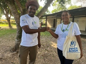 Homem negro e mulher indígena apertam as mãos, ambos olhando para a câmera. Ela carrega uma bolsa ecológica com a logomarca do ACNUR. Ao fundo existem árvores e uma casa.