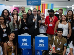 Grupo de pessoas reunidas em um estande da ONU com dois totens azuis exibindo o logotipo do ACNUR (Agência da ONU para Refugiados). Ao fundo, há um painel com faixas coloridas e outro com o emblema das Nações Unidas. Algumas pessoas usam crachás e seguram sacolas, indicando participação na COP30.