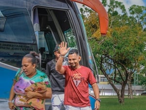 Pessoas descendo de um ônibus estacionado; uma delas acena com a mão enquanto outra caminha carregando uma criança enrolada em um cobertor. Árvores e uma área verde aparecem ao fundo.