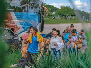 Grupo de pessoas caminhando com malas e crianças próximo a um ônibus estacionado, em uma área aberta com vegetação em primeiro plano e árvores ao fundo.
