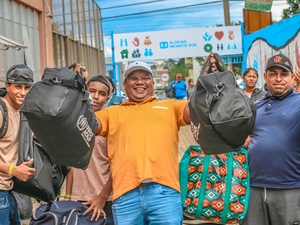 Grupo de pessoas carregando mochilas e bolsas grandes com a logomarca do ACNUR, enquanto caminham por uma área externa. Ao fundo, há um portão com a sinalização da organização ‘Aldeias Infantis SOS’. As pessoas parecem estar em movimento, algumas levantando as bagagens, com construções e um mural colorido ao redor.