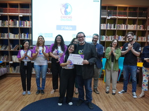 Grupo de pessoas reunidas em uma biblioteca durante um evento do programa ‘Chicas Digitais’. No centro, duas pessoas seguram um certificado. Outras pessoas ao fundo aplaudem. Atrás do grupo há uma tela de projeção exibindo o logotipo do evento e estantes cheias de livros.