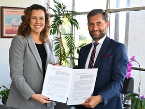 Duas pessoas em ambiente institucional seguram documento oficial aberto após assinatura, com plantas e janelas ao fundo