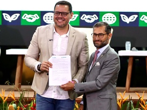 Entrega oficial de documento durante evento institucional, com participantes no palco e painel de identidade visual ao fundo.