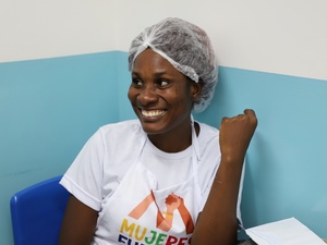 Mulher está sentada frente a uma parede azul e branca. Posa para a foto sorrindo. Ela está vestida com uma camiseta branca com a marca do projeto Mujeres Fuertes. Ela leva touca no cabelo. 