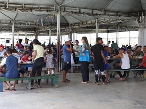 Dezenas de pessoas ocupam um amplo refeitório coberto, sentadas em longas mesas e bancos de madeira enquanto fazem refeições e conversam. Algumas pessoas circulam pelo local, que possui estrutura metálica, piso de concreto e ambiente aberto e coletivo.
