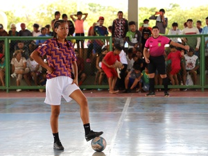 Pessoa participante de uma partida de futsal em uma quadra coberta, com um pé apoiado sobre a bola próximo à linha central. A pessoa veste camisa estampada em laranja e roxo, shorts brancos, meias pretas e chuteiras pretas. Ao fundo, atrás de uma grade verde, há um grande público acompanhando o jogo. Na lateral da quadra, outra pessoa com uniforme rosa está posicionada próxima à área de jogo. A quadra é iluminada pela luz natural, com árvores visíveis ao fundo.