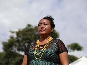 Alida usa colares de contas amarelas posa ao ar livre durante o dia. A imagem foi feita em ângulo baixo, destacando a pessoa contra o céu parcialmente nublado. Ao fundo, aparecem árvores desfocadas e parte de uma tenda, criando um contexto de evento realizado em área aberta.