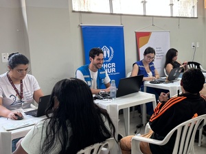 Pessoas atendem solicitantes em uma sala com mesas, computadores e cadeiras durante ação de atualização documental. Ao fundo, banners do ACNUR e do Serviço Jesuíta a Migrantes e Refugiados (SJMR) estão posicionados atrás da equipe de atendimento.
