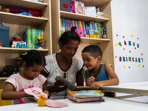 Crianças e uma pessoa adulta folheiam livros em um espaço de leitura com estantes repletas de materiais educativos e brinquedos.