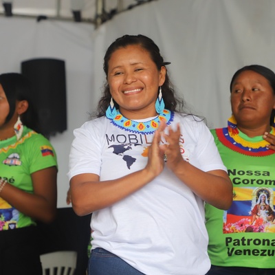 Três pessoas de pé, todas com colares coloridos. A pessoa no centro está usando uma camiseta branca com o texto 'MOBILIS' e um mapa do mundo. As duas outras pessoas estão usando camisetas verdes com a imagem de Nossa Senhora de Coromoto e o texto 'Nossa mãe Coromoto Patrona de Venezuela'
