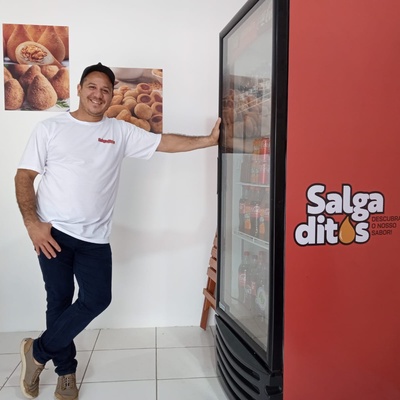 Um homem vestindo uma camiseta branca e calças escuras está de pé com uma mão apoiada em uma máquina de venda automática vermelha. A máquina tem o texto "Salgaditos: Descubra o Melhor Sabor" escrito nela. Há dois cartazes na parede atrás dele, ambos exibindo imagens de salgados. O chão é de azulejos e há uma porta fechada ao fundo.