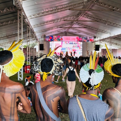 Um grupo de quatro indígenas com pintura corporal tradicional e cocares de penas está de costas para a câmera, voltado para um palco sob uma grande tenda. O palco está bem iluminado e apresenta artistas, enquanto uma plateia assiste.