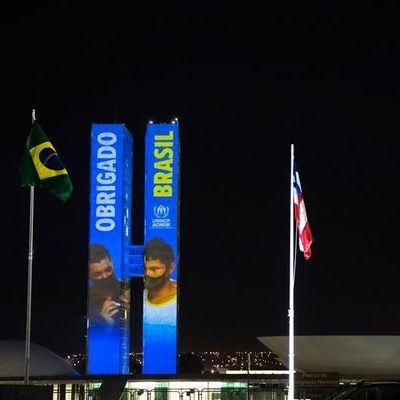 As duas torres do Congresso Nacional do Brasil estão iluminadas à noite e exibem as palavras 'OBRIGADO BRASIL' em letras grandes, com 'OBRIGADO' em azul no prédio da esquerda e 'BRASIL' em amarelo no da direita. Imagens de pessoas são projetadas nas fachadas. Em frente, há dois mastros com bandeiras — a do Brasil à esquerda e outra não identificada à direita. O céu está escuro e há luzes da cidade ao fundo."
