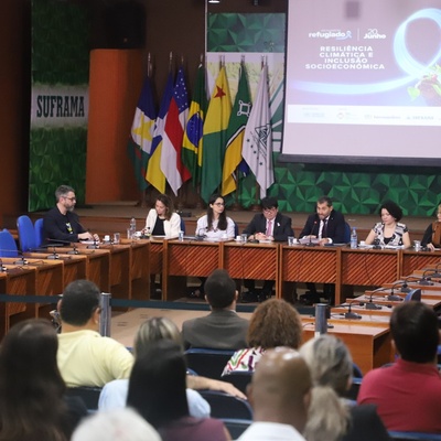 Evento oficial com várias pessoas sentadas em uma mesa longa à frente de um auditório. Atrás dos palestrantes, há bandeiras do Brasil e de estados brasileiros. Um telão exibe a frase 'resiliência climática e inclusão socioeconômica'. A palavra 'SUFRAMA' aparece à esquerda, indicando relação com a Superintendência da Zona Franca de Manaus.