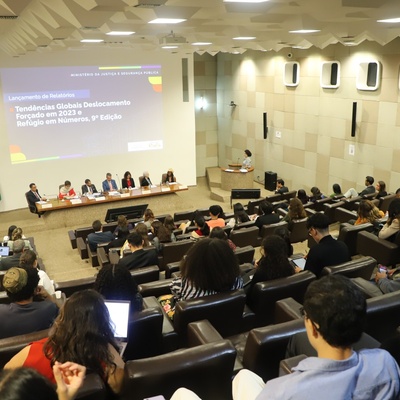 Pessoas em um auditório assistindo a outras pessoas em uma mesa com uma grande tela ao fundo. 