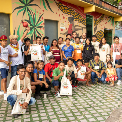 Grupo de pessoas posando em frente a um mural colorido com desenhos abstratos e a figura de uma pessoa segurando um objeto. 