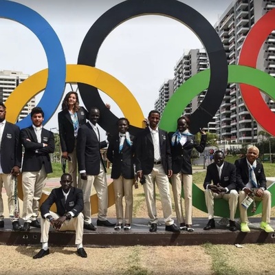 Grupo de pessoas vestidas com trajes formais posando em frente à escultura dos anéis olímpicos, com prédios altos e céu claro ao fundo.