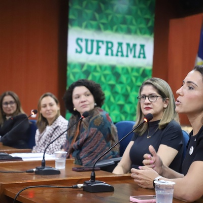 Painel com seis mulheres sentadas à mesa com microfones, participando de uma conferência. Ao fundo, há uma faixa verde e branca com o texto 'SUFRAMA'. Sobre a mesa, há garrafas de água e copos.