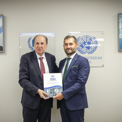 Dois homens de terno posam diante de uma parede com dois retratos emoldurados e dois logotipos das Nações Unidas, segurando um documento intitulado 'UNIDADES ACADÊMICAS'.