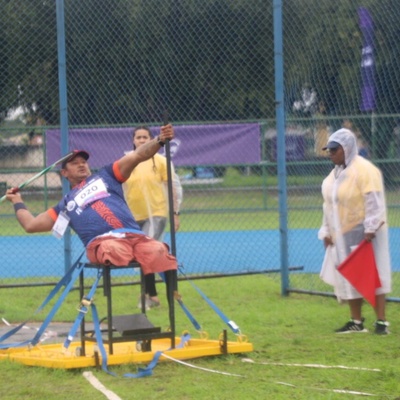 Atleta com uniforme azul e vermelho, número 070 no peito, se prepara para lançar dardo em uma plataforma amarela. Duas pessoas estão próximas, uma segurando uma bandeira vermelha. Ao fundo, há uma cerca, árvores e estruturas desfocadas.