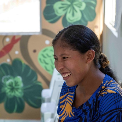mulher indígena de perfil com camiseta azul e pinturas de flores verdes ao fundo.