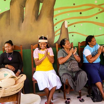 Grupo de pessoas indígenas sentadas em uma fileira de cadeiras, algumas aplaudindo e sorrindo, em frente a um mural colorido com arte representando uma mão em destaque e elementos gráficos abstratos. Cestos artesanais indígenas estão sobre uma mesa à esquerda.