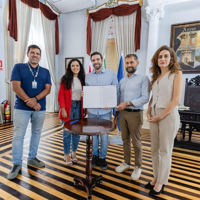 Cinco pessoas em pé em uma sala decorada com janelas grandes, cortinas, uma pintura ao fundo e uma mesa de madeira ao centro. Duas delas seguram um livro ou documento aberto. O chão tem padrão listrado.