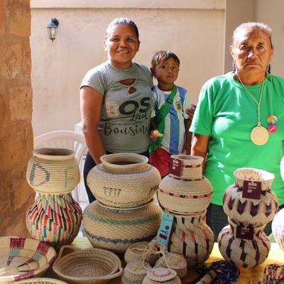 Três pessoas indígenas em pé, sendo duas mulheres e uma criança ao meio, atrás de uma mesa com cestos e itens artesanais de diferentes formas, tamanhos e cores, exibindo padrões detalhados. 