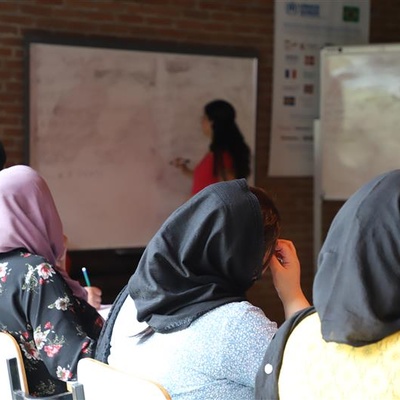 Sala de aula com estudantes sentadas, usando lenços na cabeça, voltadas para um quadro branco onde uma pessoa instrutora escreve ou explica algo. Ao lado do quadro há um flip chart, e ao fundo, uma parede de tijolos com cartazes.